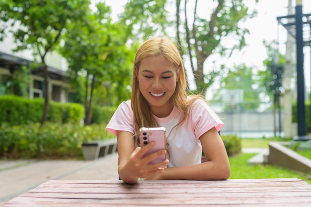 Портрет красивой азиатской девушки с светлыми волосами, улыбающейся, используя телефон на открытом воздухе, сидя.