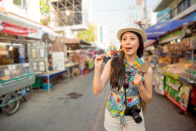 アジアの休暇旅行で、タイの地元のフードストリートマーケットを歩いて、おいしい焼けた鶏肉のケバブを味わう美しいアジアの観光客のポートレート。