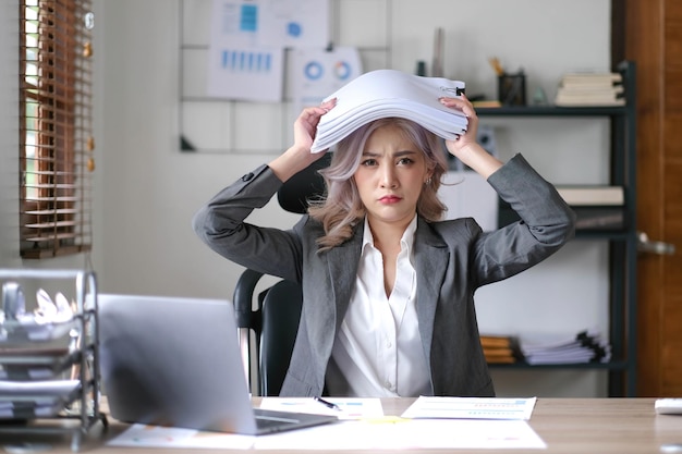 Portrait of beautiful asian woman at the desk with paperwork on her head business overwhelmed stressed concept