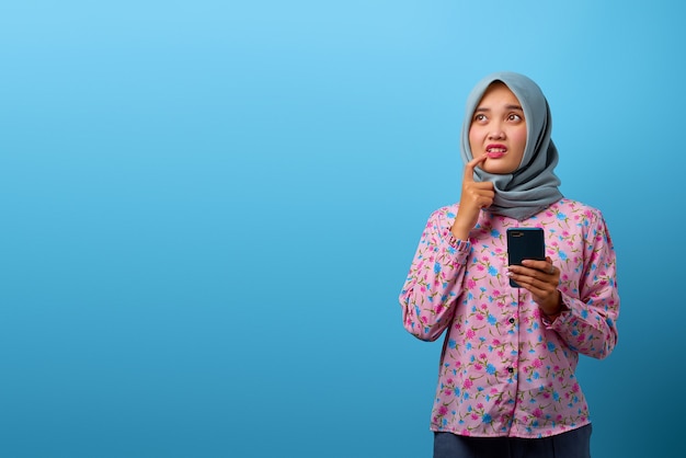Портрет красивой азиатской женщины, держащей мобильный телефон и думающей о чем-то с пальцем на подбородке
