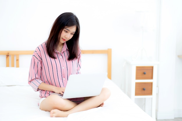 Ritratto bella donna asiatica che si siede sul letto utilizzando il computer portatile