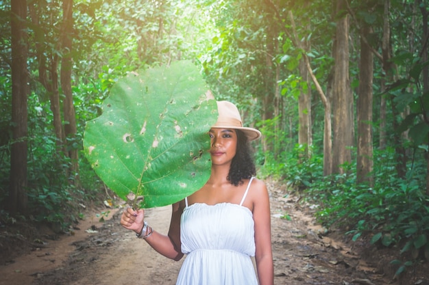 Portrait of beautiful Asian woman standing with big tree’s leaf cover half of her at forest