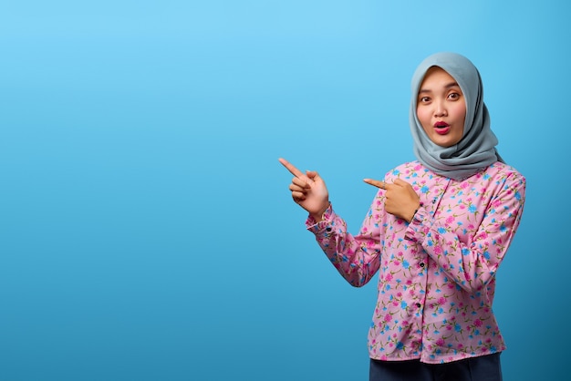 Portrait of beautiful Asian woman surprised and pointing to copy space on blue background