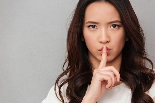 Portrait of beautiful asian woman with brown hair holding index finger on lips asking to keep silence, over gray wall