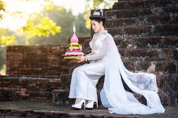 Portrait Beautiful asian woman with Thai white traditional dress costume holding Krathong and sitting in front of Pagoda temple at the ancient city ThailandLoy Krathong FestivalTransgender model