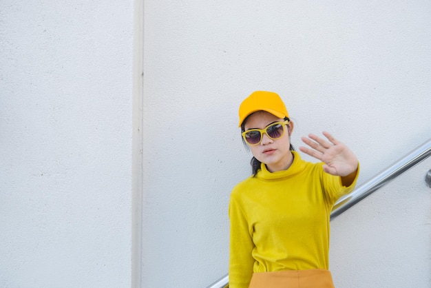 노란색 옷을 입은 아름다운 아시아 여성의 초상화는 손을 멈추게 합니다.Hipsters 소녀는 사진을 찍기 위해 계단에서 노란색 모자를 쓰고 있습니다.