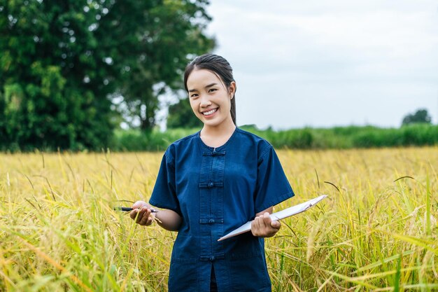 Portrait beautiful Asian young woman farmer use clipboard checking during work in organic rice field and smile with happiness