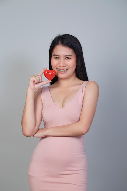 Portrait of beautiful Asian young woman holding pointing red heart shape isolated on light gray background with copy space