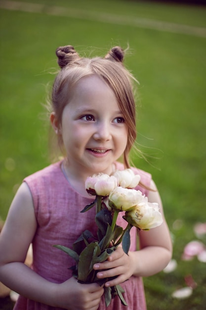 Portrait of a beautiful baby girl in a pink dress standing with a bouquet of flowers pions on a green meadow in summer