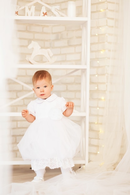 Portrait of a beautiful baby girl in a white dress in the interior with Christmas decorations