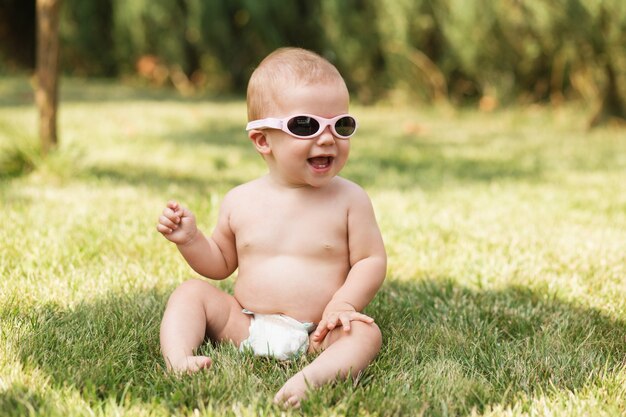 Portrait of a beautiful baby wearing diaper sitting on green grass outdoors on hot summer day