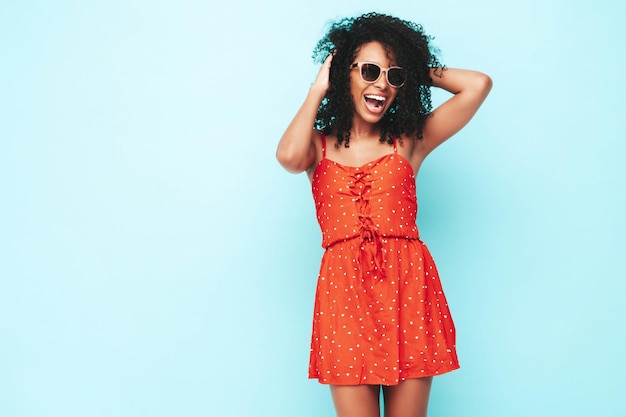 Portrait of beautiful black woman with afro curls hairstyle Smiling model dressed in red summer dress Sexy carefree female posing near blue wall in studio Tanned and cheerful In sunglasses