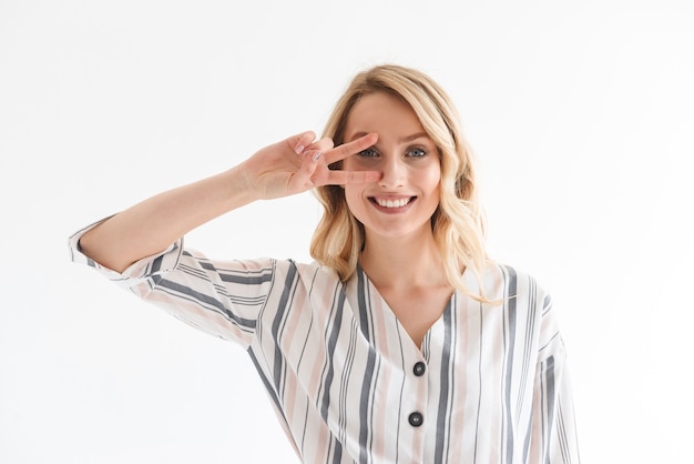 Portrait of beautiful blond woman 20s wearing casual clothes smiling at camera while showing peace sing isolated over white wall