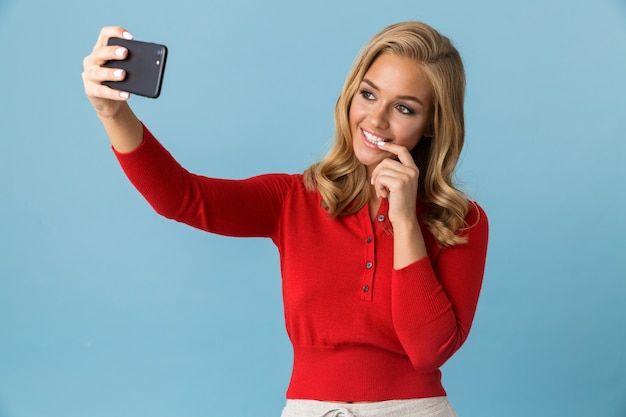 웃 고 파란색 벽에 고립 된 휴대 전화에 셀카 사진을 찍고 빨간 셔츠를 입고 아름 다운 금발 여자 20 대의 초상화