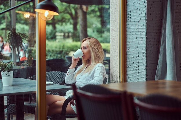 Portrait of a beautiful blonde female dressed in a white blouse sitting at a table drinks coffee at terrace cafe.