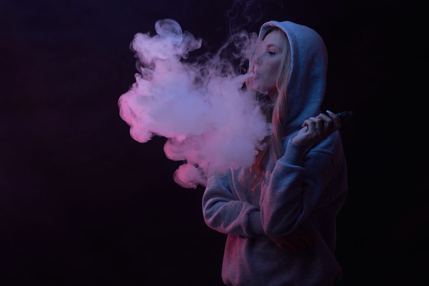 Портрет красивой блондинки в серой толстовке с капюшоном курит вейп на черном фоне студии, облако парового дыма, мини-кальян