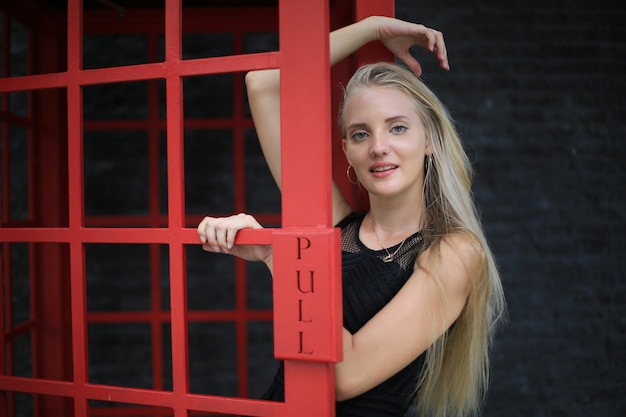 Portrait of Beautiful blonde hair girl on black dress standing in red phone booth 