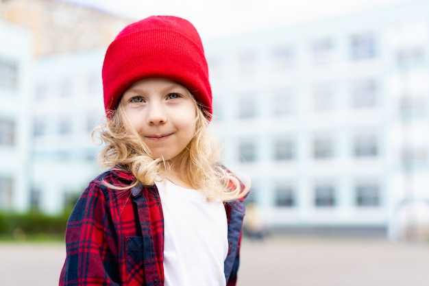 Portrait of a beautiful blonde little girl on the street, wearing a red hat and a shirt in a cage