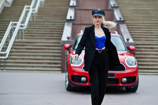 Portrait of beautiful blonde sexy fashion woman model in cap and in all black blue corset with bright makeup near red city car