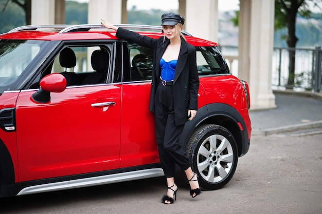 Photo portrait of beautiful blonde sexy fashion woman model in cap and in all black blue corset with bright makeup near red city car