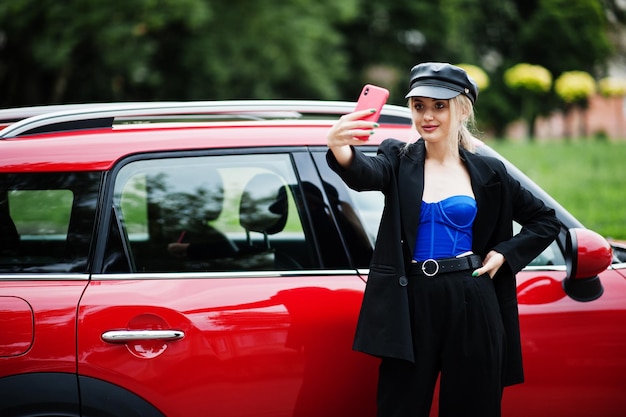 Ritratto di bella bionda modello di donna di moda sexy in berretto e in tutto nero con trucco luminoso vicino a un'auto da città rossa con il telefono cellulare