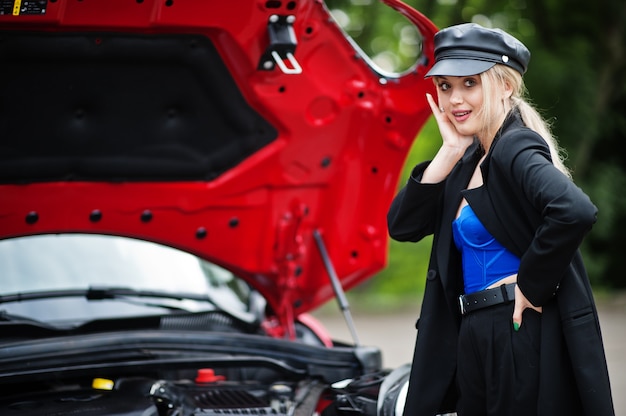 Ritratto di bella modella bionda sexy moda donna in berretto e tutto nero con trucco luminoso vicino a city car rossa con cappuccio aperto.