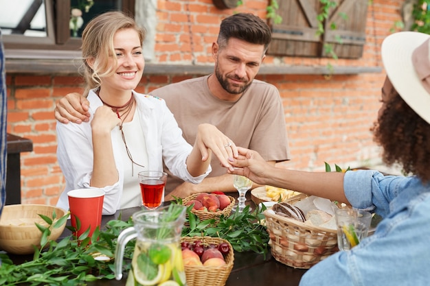 Portrait of beautiful blonde woman showing off engagement ring to friends during outdoor dinner