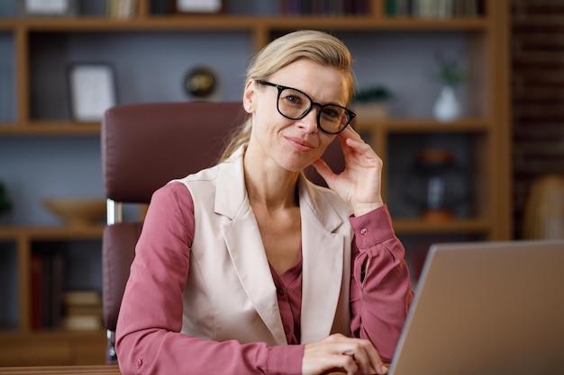 카메라를 보고 웃 고 아름 다운 금발 여자의 초상화 사무실에서 직장에 앉아 안경을 착용 하는 성인 세련 된 자신감 사업가 현대 성공적인 여성 지도자의 초상화