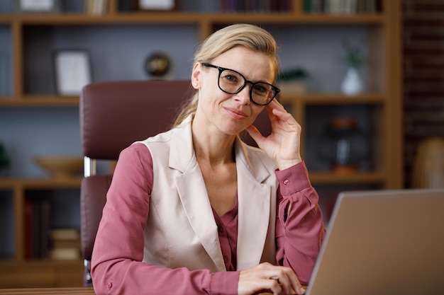 Foto ritratto di bella donna bionda che guarda l'obbiettivo e sorridente donna d'affari sicura di sé alla moda adulta che indossa occhiali seduti sul posto di lavoro in ufficio ritratto di moderna leader femminile di successo