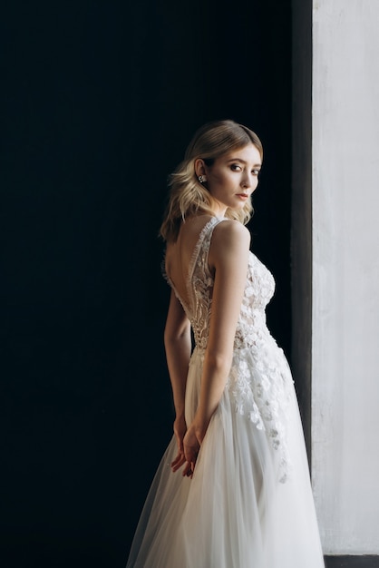 Portrait of beautiful bride with blonde hair in a white dress on the black background. bride holds tender hands behind her back. copy space