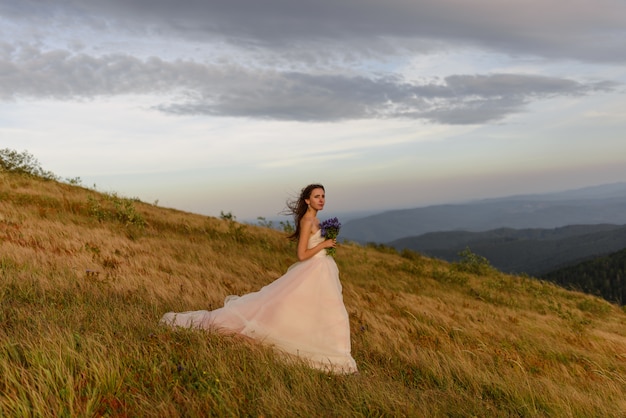 Portrait of a beautiful bride with a bouquet of wildflowers "Cornflowers" on a landscape of autumn mountains. The wind blows her hair. Wedding ceremony on top of the mountain.