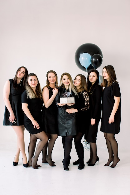 Portrait of beautiful bridesmaids in black dresses with attractive bride-to-be in the middle holding delicious cake. Background is decorated with air balloons.