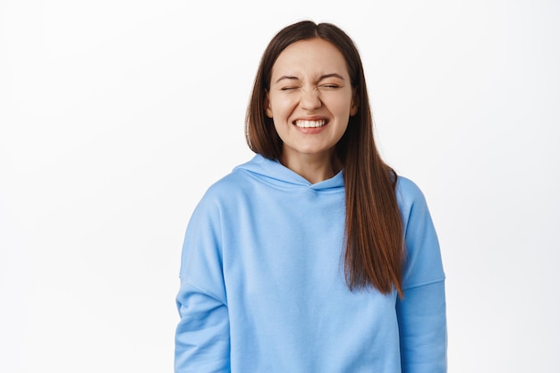 Portrait of beautiful brunette girl close eyes, smiling carefree, imaging, dreaming of something, grinning from delight, standing in hoodie against white background