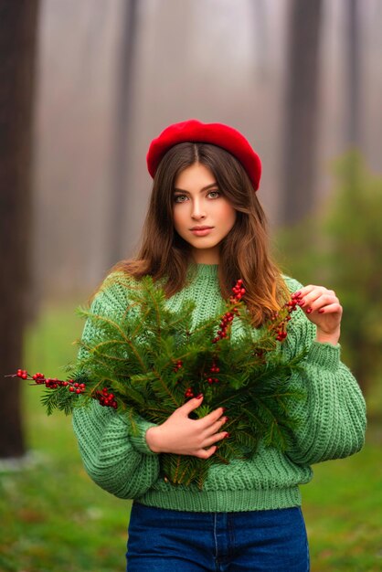 Портрет красивой брюнетки в красной шляпе и зеленом вязаном свитере с рождественскими ветвями