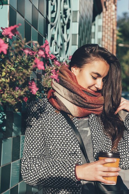 路上でテイクアウトドリンクと美しいブルネットの少女の肖像画。市内のスカーフと秋のコート。街の通りを歩いてコーヒーを飲みながらビジネスレディの素晴らしい景色。