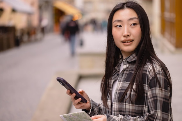 Photo portrait of a beautiful brunette korean woman holding a map and comparing it with smartphone navigation on the streets of the old city asian tourist woman traveling in europe