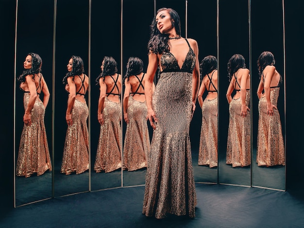 鏡の近くに立っている黄金のドレスの美しいブルネットの女性の肖像画ファッションスタイル