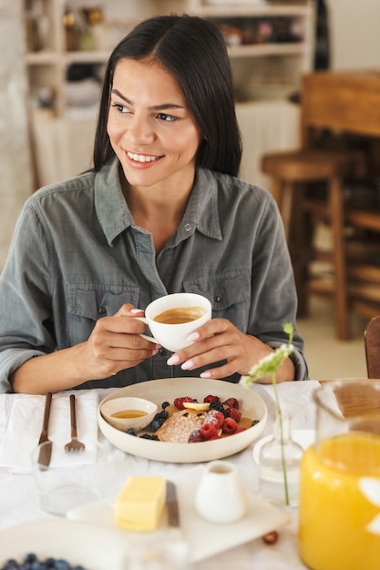 Photo portrait of beautiful brunette woman sitting at table in bright kitchen while having breakfast at home in morning