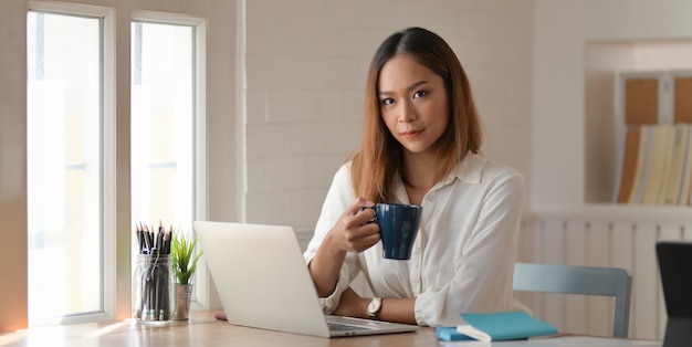 Ritratto di bella imprenditrice bere caffè mentre si lavora in ufficio e sorridendo alla telecamera