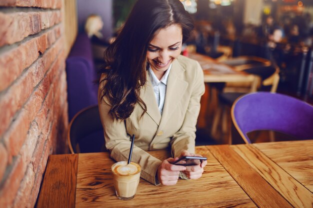 美しい白人ブルネットの肖像画は、カフェに座っているスマートカジュアルな服を着て、メッセージを送信するためにスマートフォンを使用しています。木製のテーブルには、新鮮でおいしいコーヒーがあります。