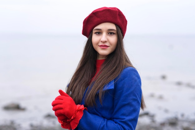 Portrait of beautiful Caucasian girl, young attractive stylish elegant brunette woman with long hair in red beret and gloves and blue coat looking at camera outdoors, walking on beach, ocean.