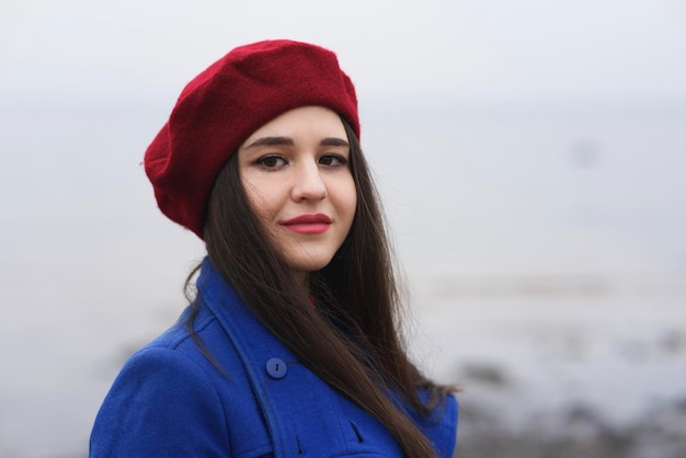 Portrait of beautiful Caucasian girl, young attractive stylish elegant brunette woman with long hair in red beret and gloves and blue coat looking at camera outdoors, walking on beach, ocean.