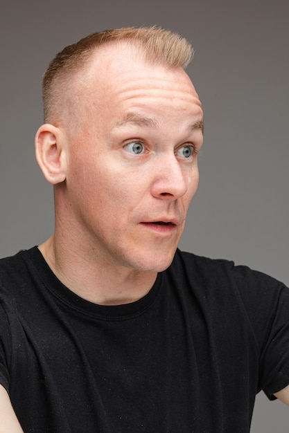 Portrait of beautiful caucasian male in black t-shirt looks at something interesting with big eyes and open mouth isolated on grey background