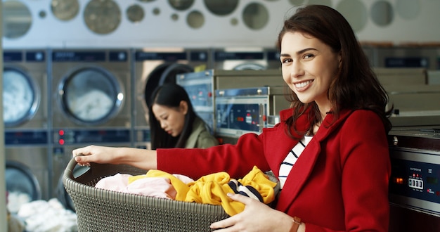 Photo portrait of beautiful caucasian stylish woman smiling to camera and holding basket with dirty clothes while standing in laundry service. pretty girl laughing clean clothing at washing machines.