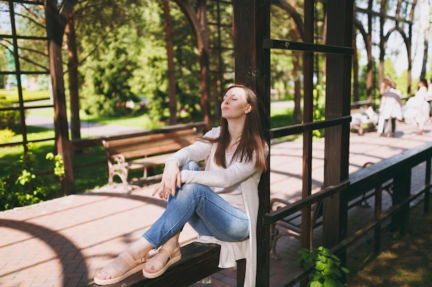 Portrait of beautiful caucasian young woman with charming smile wearing light casual clothes. Smiling female sitting under archway in city park in street outdoors on spring nature. Lifestyle concept.