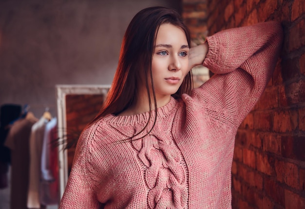 Portrait of a beautiful charming brunette dressed in stylish clothes leaning against a brick wall in a room with loft interior.