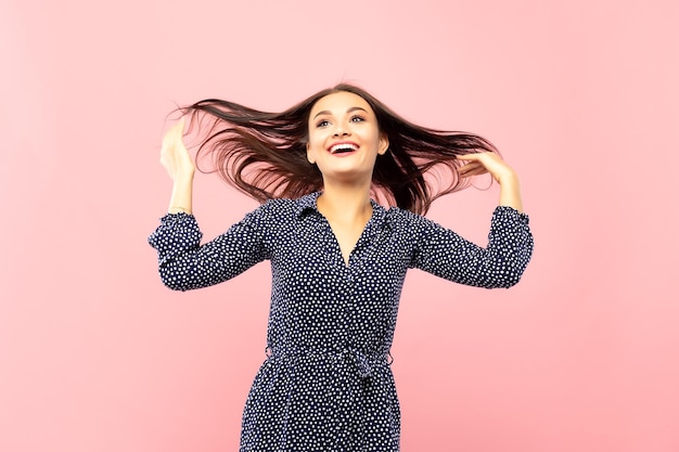 Portrait of a beautiful cheerful brunette in a blue dress with white polka dots with flying dark hair smiling laughing looking at the camera on a pink background