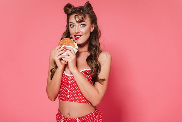 Portrait of a beautiful cheerful pin-up woman wearing swimsuit standing isolated, eating a burger