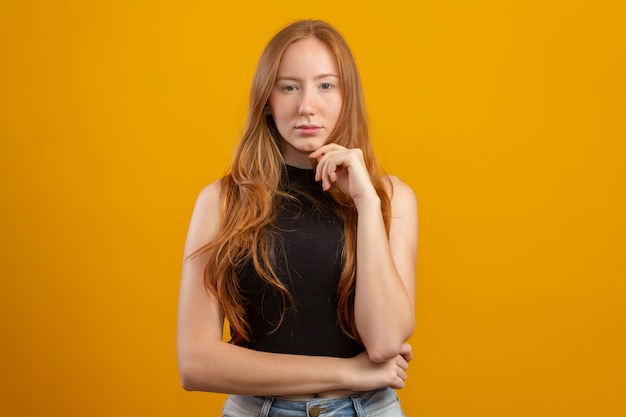 Portrait of beautiful cheerful redhead girl with a hand on the chin  over yellow wall