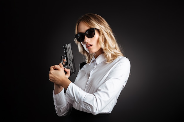 Portrait of a beautiful confident blonde haired woman dressed in formal clothes standing isolated over black wall, wearing sunglasses and carrying gun